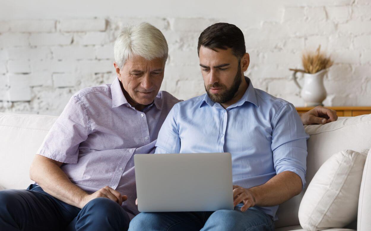 father and son sit together on a couch reviewing the cyber security effectiveness on the father’s laptop taking action to minimise his online risk
