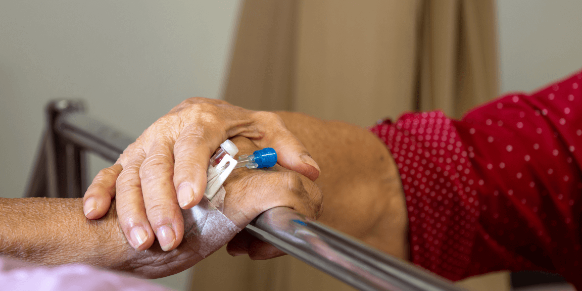 older woman's hand on husband's hand as she comforts him in his hospital bed: canula showing: she contemplates their future. seeking a better aged care outcome