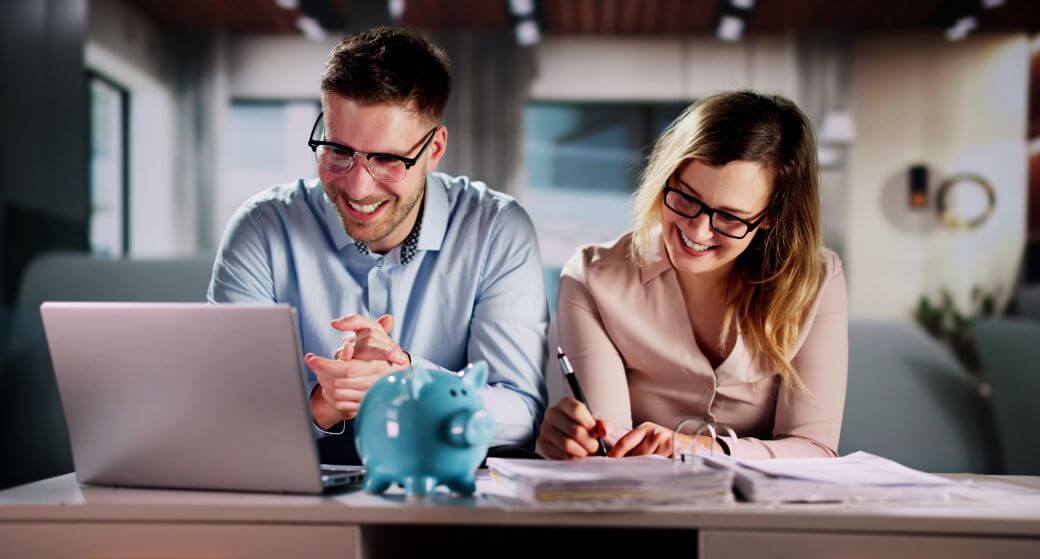 mature aged couple delighted with the retirement plan they envisaged and now formalised with their financial adviser as they sit at their desk with the plan open on their laptop, a folder of supporting documents open, and a blue piggy bank reflecting a savings strategy