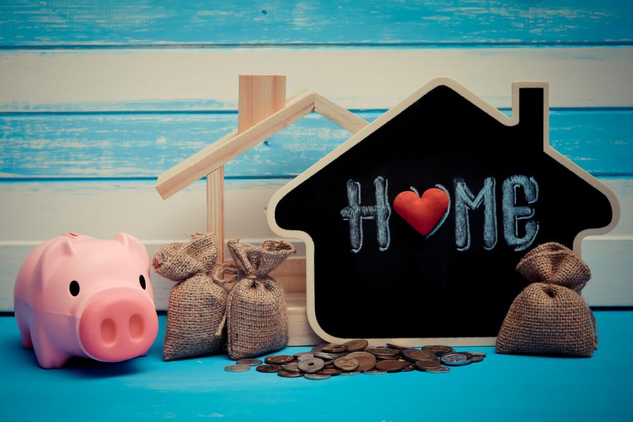 image shows an outline of a house (home) together with a pink piggy bank, some hessian money sacks and some coins on a blue table surface and against a blue and white horizontal striped background; together indicating saving to accumulate a home deposit