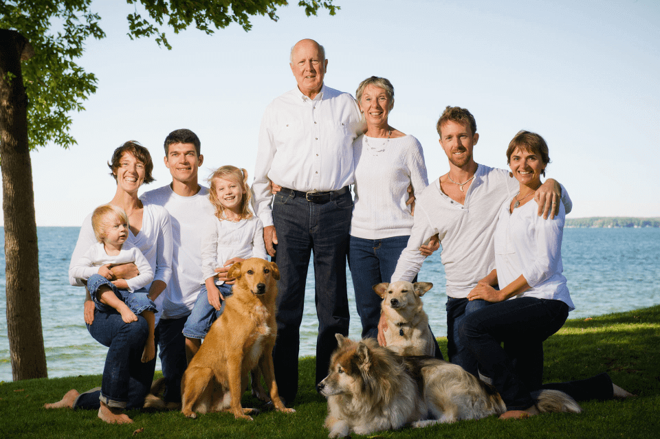 three generations of a family - grandparents, their children, spouses and grandchildren posing with the three family pet dogs in the shade of a tree with a calm water bay background - having discussed superannuation death benefits and business succession planning, including key-man insurance