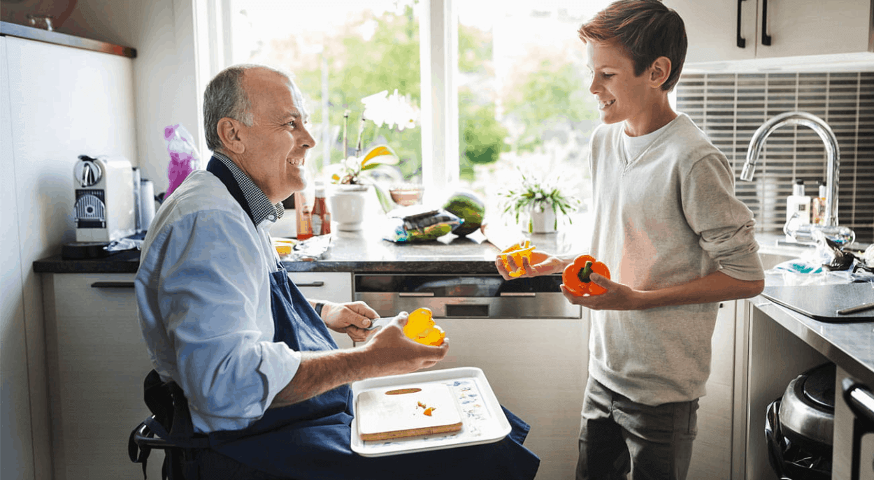 a wheelchair-bound older man talks to young boy, possibly his grandson in his kitchen, potentially discussing how income protection insurance preserves his wealth and lifestyle