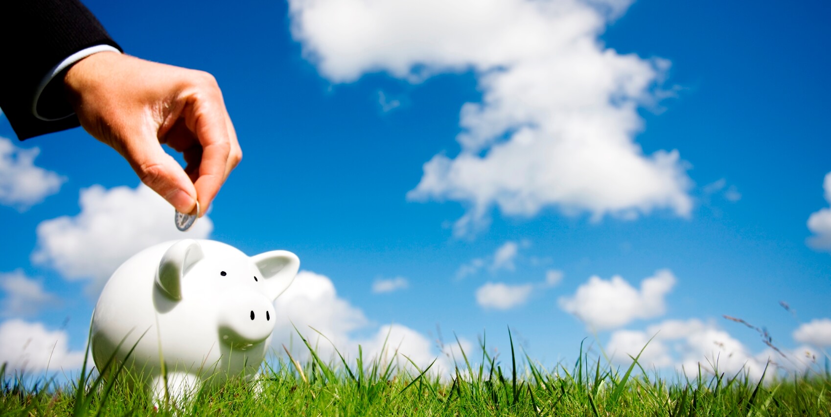 a hand shown dropping a coin into a white piggybank against a lightly clouded blue sky and sitting on fresh green grass - representing making a personal superannuation contribution, an eligible government co-contribution, or an employer default fund chosen by employee