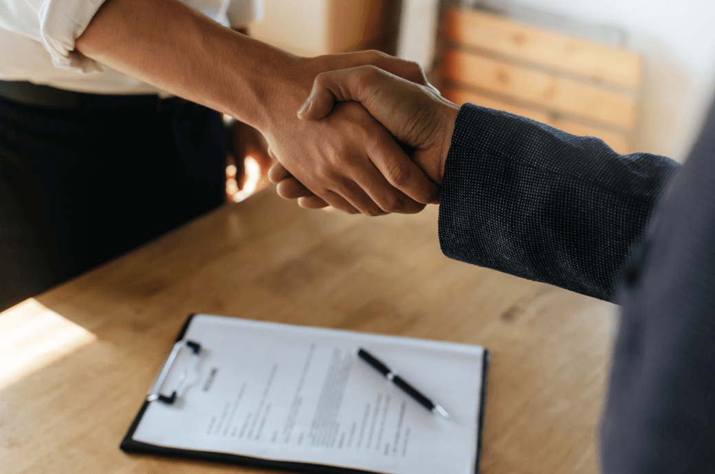 men standing across a desk shaking hands after signing a business agreement that sits on the desk below their hands - business succession strategy agreement