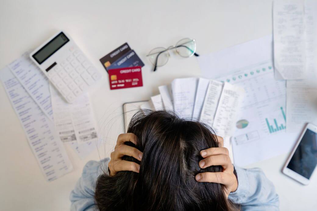 looking down onto the head in hands of a young female stressing over her financial affairs with credit cards, purchase invoices, calculator , smart phone and charts strewn across the desk in front of her - credit card debt management inhibiting her financial well-being - and her busy-ness frustrating attempts to secure life insurance