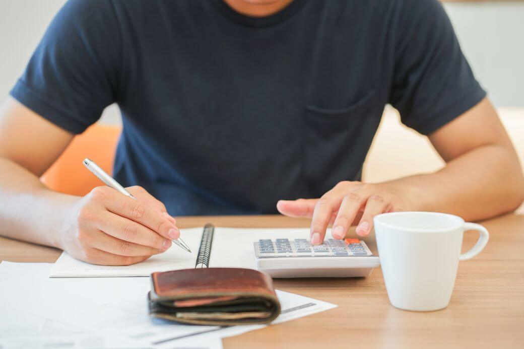 mature young man sitting at desk with notepad and calculator ready, wallet in foreground and coffee cup to his side, analysing financial position with view to debt management