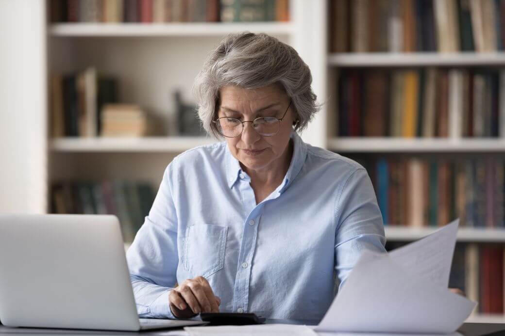 woman working in home office wearing a blue cotton shirt, sitting at her desk with an open laptop, using a calculator, and reading document - perhaps reviewing the investment performance of her SMSF portfolio compared to the investment strategy; she may well have had some Estate Planning prompts
