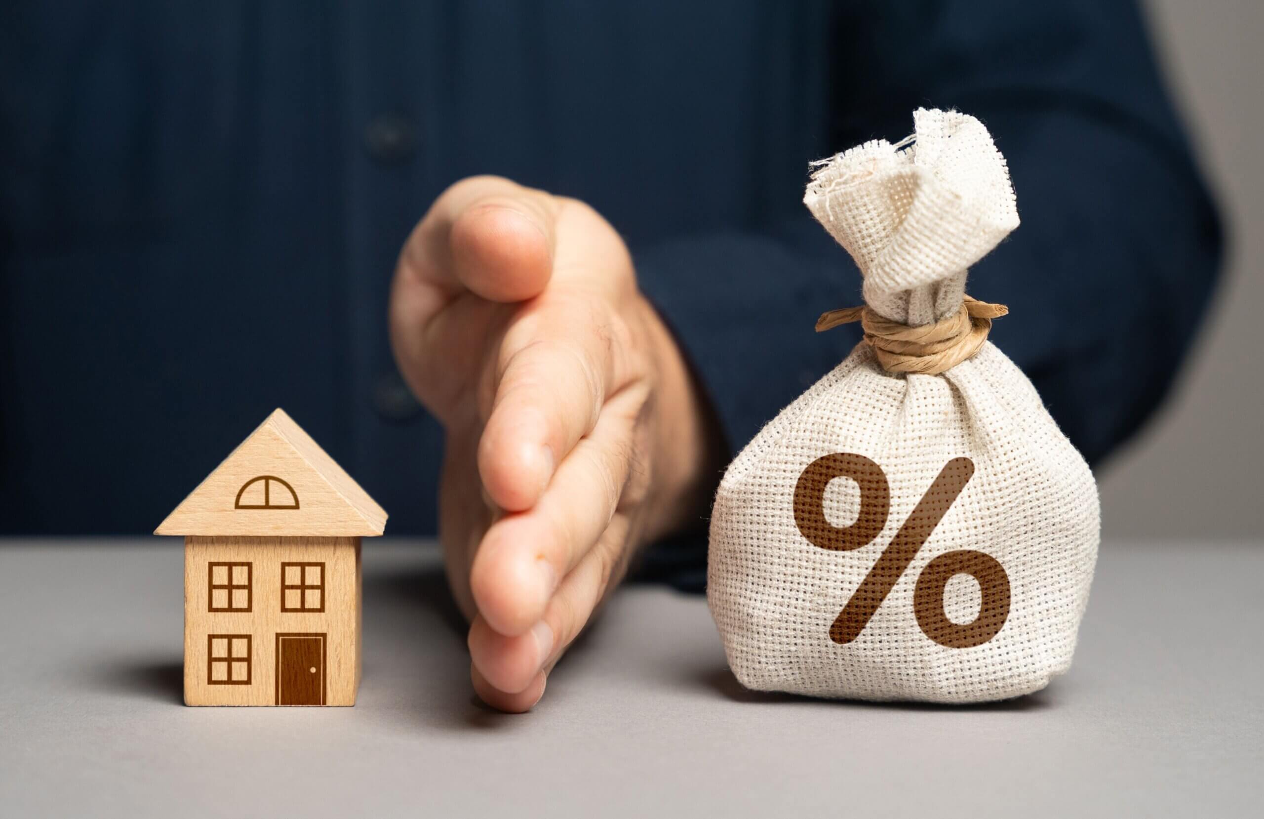 a man's hand is on edge on the table forming a barrier between the home he has mortgaged and the interest that accrues against that mortgage - he has a fixed interest rate contract