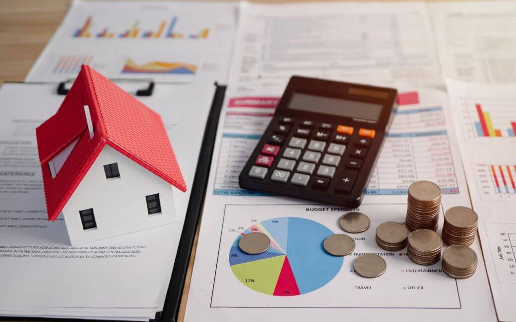 documents, coinage, a calculator, and a tiny model house pread across a desk top and representing investment in mortgages under a mutual fund trust