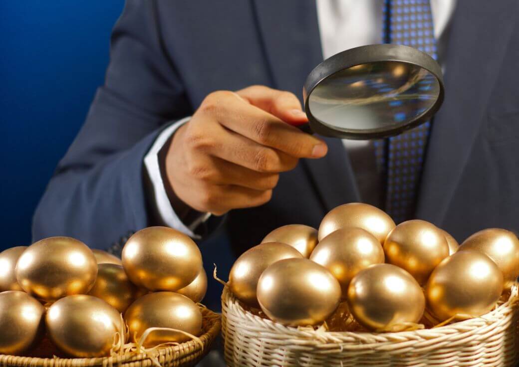 a man dressed in a suit holds a magnifying glass over a pair of baskets of golden eggs that depict the wealth accumulation he is achievinmg through superannuation contributions