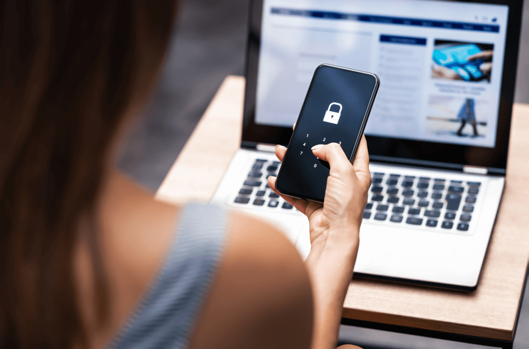 young woman faces her mobile device and a laptop, exercising good cyber security practice using multi-factor authorisation to securely access her online accounts