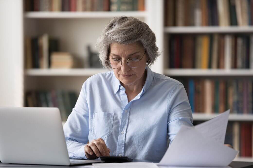 woman sitting in home office reviewing investment progress on her laptop, using calculator and reading financial report on the Child Endowment Bond she has for her granddaughter