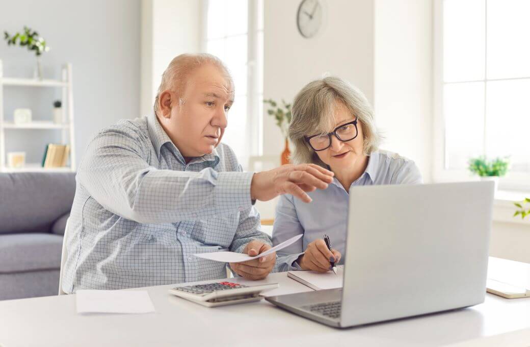 older couple sitting at home table reviewing finances and retirement funds and using a calculator and a laptop to calculate allocated pensions