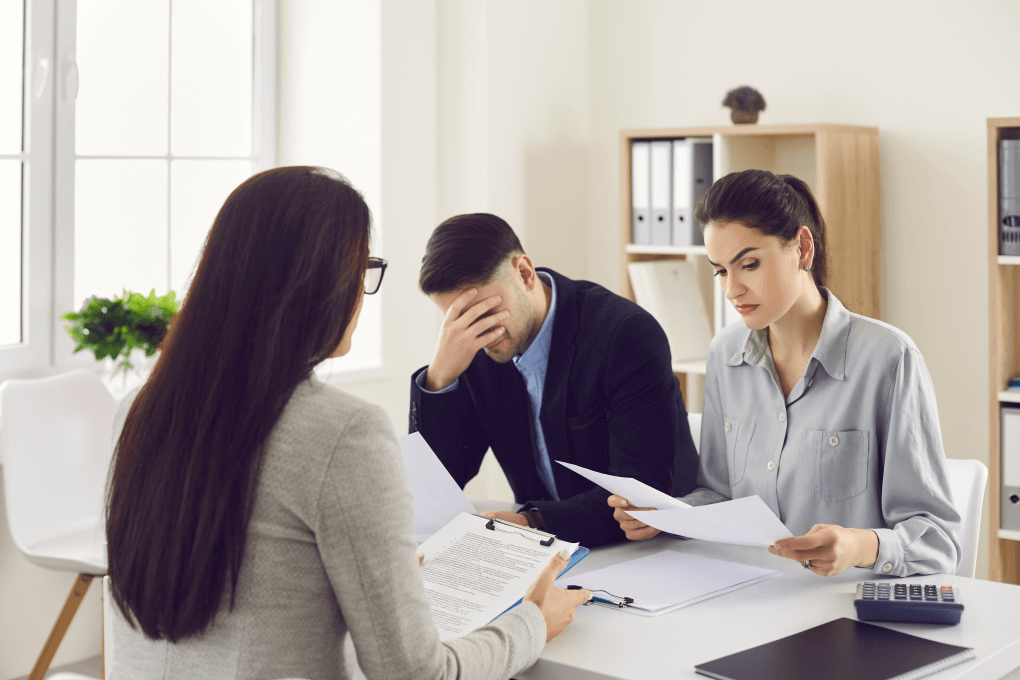 young couple duped by a scammer getting news from adviser that their money is lost as they sit dejected across the desk from her - demonstrating investment returns promised attract but the outcome is tragic