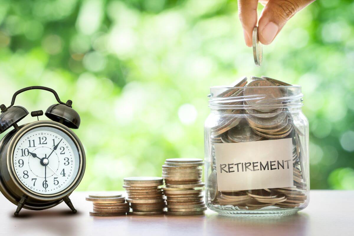 analogue clock and a jar to which money is being added, book-end three stacks of coins: they sit on a table in a sunny garden setting. The jar has the word Retirement on a label accumulating funding for the saver’s dream retirement