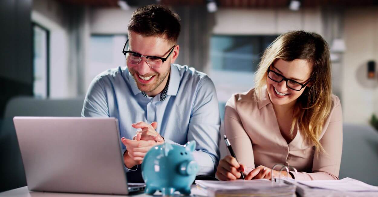 a young couple with smiling faces resolving to purposefully utilise their tax refund to achieve their longer-term financial goals as they sit in front of their computer screen and contemplate documents such as investment strategies and debt management