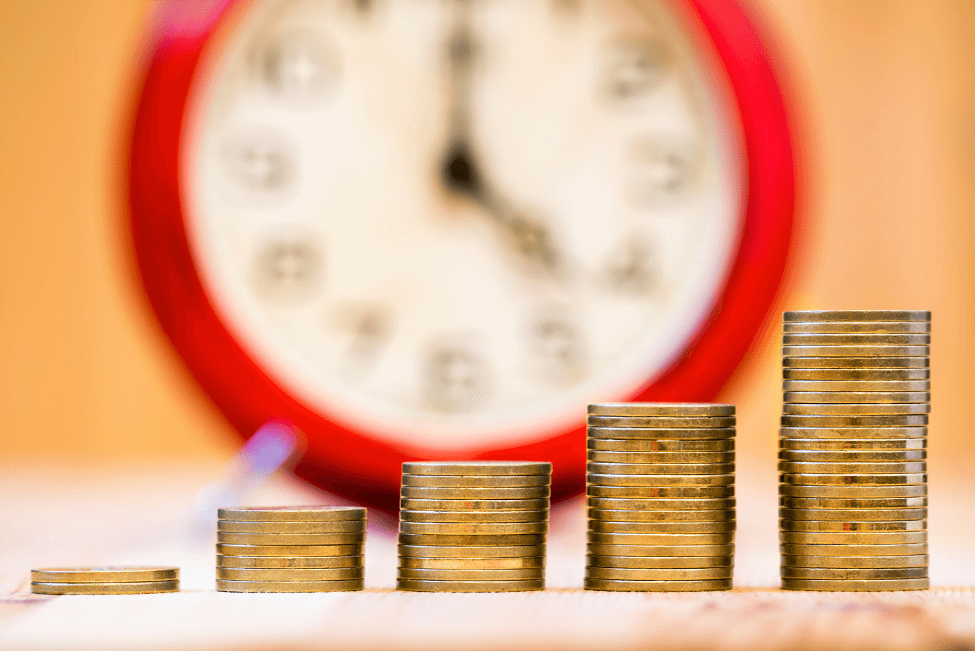 an image of a red-framed clock blurred in the background with five coin stacks focused in the forefront: from left to right each stack of coins is higher than the stack to its left showing the magic of compounding