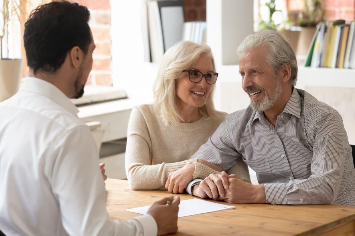 financial planner with back to camera faces a client couple sitting opposite at a small desk to present their insurance review