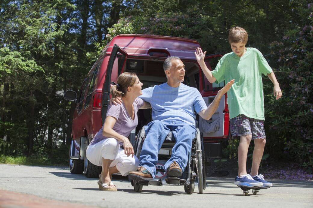 casually dressed family including a man in his wheelchair, accompanied by wife, and about to low-five with their son on his skateboard: their red van is in the background, as they celebrate that income protection insurance has enabled them to continue doing normal activities of life