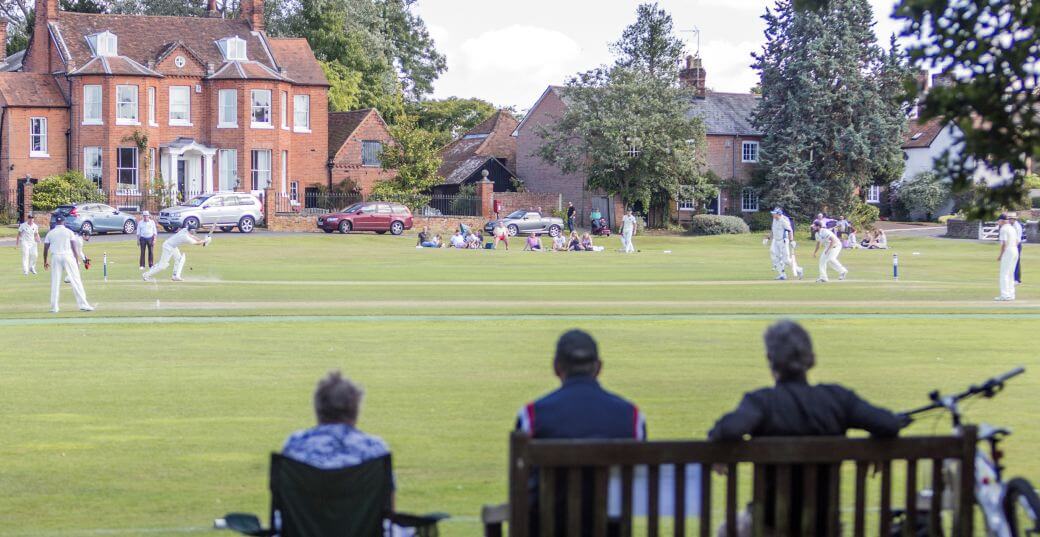 teams of cricketers in an English country setting being watched by local people supporting them on a fin, sunny day - the opening batsman had previously experienced a stroke trauma