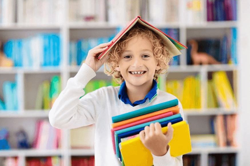 child in school classroom-library: he is wearing a blue polo shirt and a white pullover - and holding a selection of books in his left arm, and one book, opened as a shelter over his head, in his right hand. Education funding in place for this one.