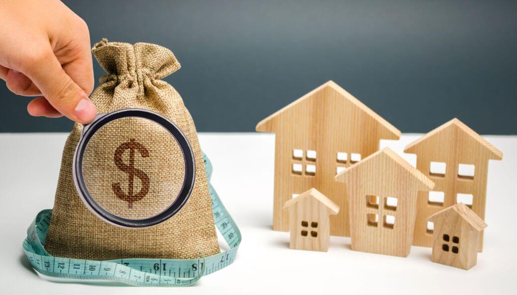 a hand holds a magnifying glass up to a dollar sign on a hessian moneysack that is encircled by a tape measure as it sits on a white table alongside some wooden block houses - signifying strategic wealth management intent