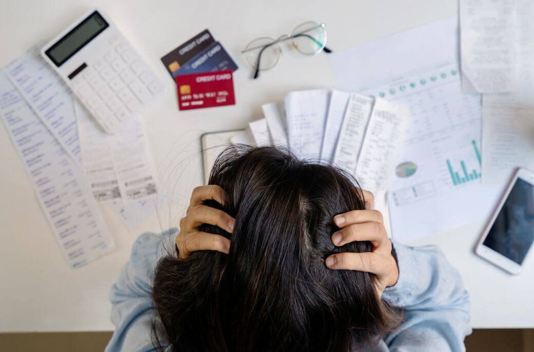 young woman despoiring as she clutches her scalp, holding her head in her hands looking over various credit card and other accounts on the desk she is leaning on - showing interest rate interest wanting to better manage her finances