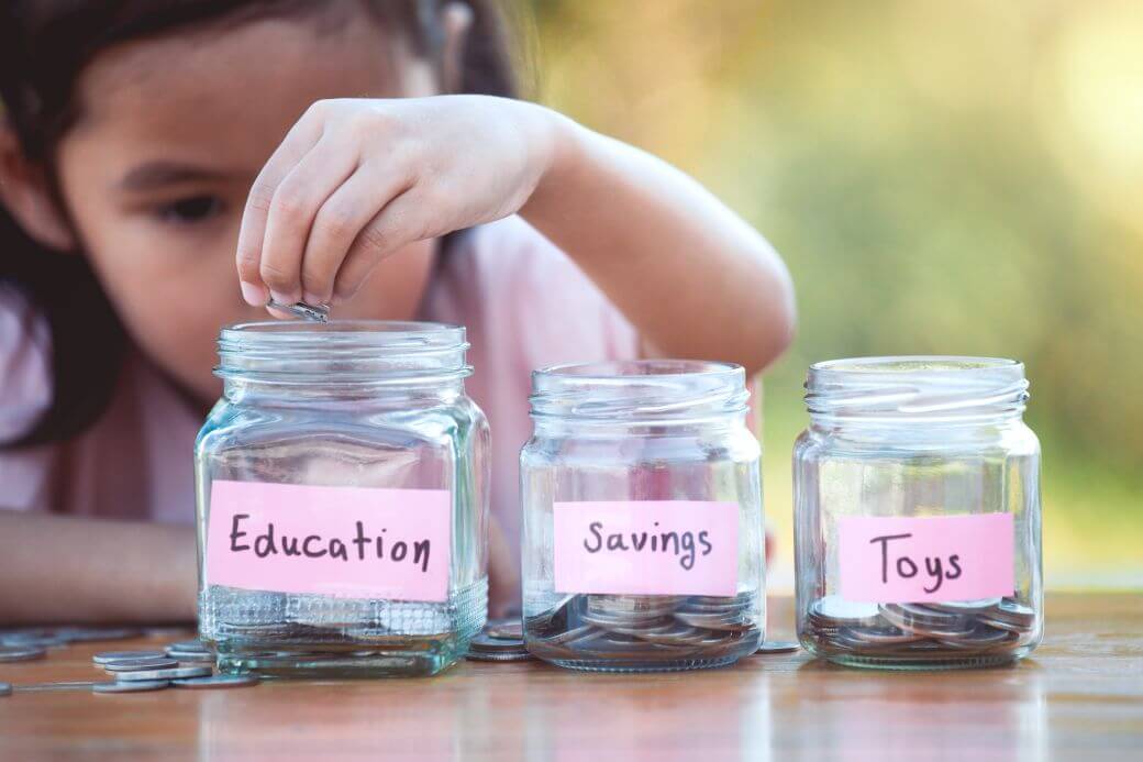 young girl diligently placing coins in jars set aside for 'education', 'savings', and 'toys' each jar labelled with pink labels. Some unallocated coins still lie on the table on which she is 'working'. Setting a strategy for funding education costs