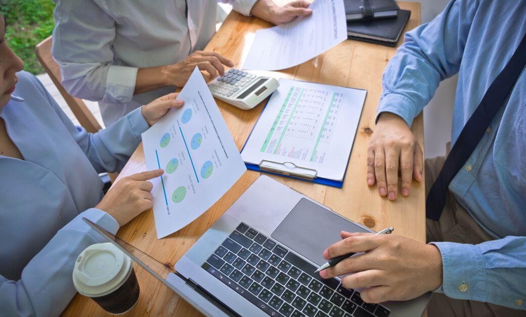 an investment team sitting around a bench with a laptop, calculators and printed reports, setting investment strategy for a client to consistently apply