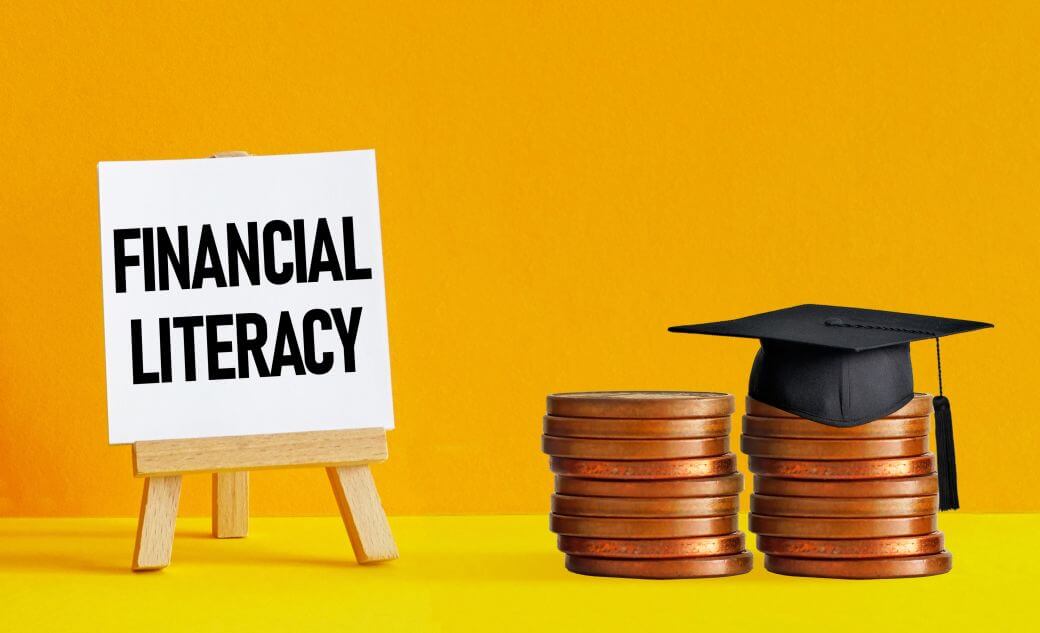 against an orange coloured background and on a yellow surface stand a signboard with nthe words financial literacy alongside two piles of coins , one with a graduates mortice board - suggesting that childrens savings account lessons have been well learned
