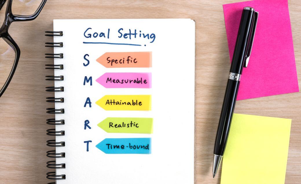 Some workshopping planning tools preparation for some goal-setting: spectacles, a notepad, a couple of post-it note stickers and a pen showing that SMART Goal Setting is happening